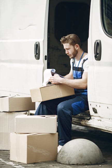 A young man with short brown hair and a beard, dressed in a striped t-shirt and blue overalls, is sitting on the edge of an open white van during a home relocation process. He is holding a clipboard o