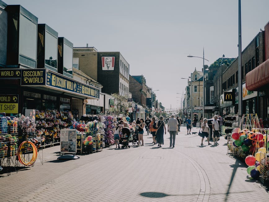 A busy street scene on Electric Avenue in Brixton with a mix of pedestrians, including individuals pushing strollers and carrying shopping bags, walking along the pavement lined with shops and market stalls. The stalls display various goods, such as flowers, balloons, and household items, with some merchandise arranged on tables and in baskets. Storefronts include a Discount World shop with a bright sign, a KFC outlet with its logo visible, and other retail units with large windows and awnings. The street is well-lit by tall street lamps, and the sky above is clear, indicating daytime. Man and a Van Brixton's removals service is relevant to this scene, as it captures an active shopping and moving environment, where household furniture, boxes, and packaging materials might be involved in a home relocation or furniture transport process. The environment suggests a vibrant urban setting, where busy movement and logistics are integral to the area's commercial activity.