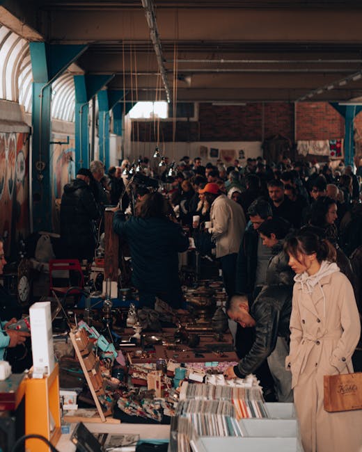 The image depicts a busy indoor market or thrift store setting with numerous shoppers browsing and examining various items displayed on long tables. The store features high ceilings made of wood or metal beams, with large windows allowing natural light to illuminate the space. Shelves and display units are visible, holding an assortment of objects such as books, vintage decorations, small appliances, and miscellaneous household items. Several customers are seen actively engaging with the merchandise, some leaning over tables to inspect items closely, while others stand nearby waiting or observing. In the background, a variety of people populate the space, creating a lively atmosphere typical of market or second-hand shops. The scene is representative of a market environment where individuals may be involved in home relocation or moving activities, with [COMPANY_NAME] potentially providing packing and moving services in the area, as suggested by the context of the [PAGE_TITLE] about market traders in Brixton.